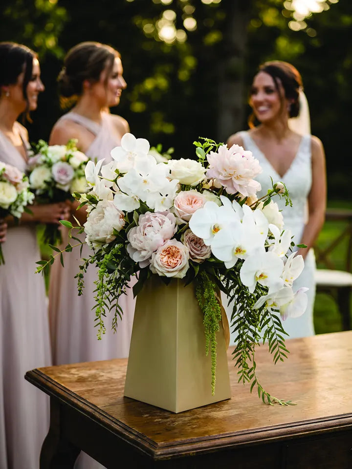 Bridal bouquet of white orchids and blush flowers in a FlowerBox, with the bridal party in the background